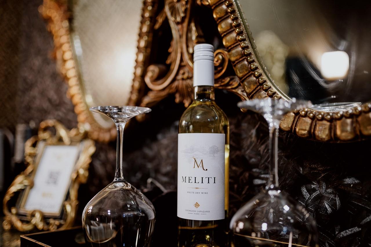 A bottle of Meliti white wine and two glasses by a gilded mirror